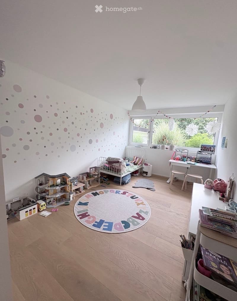 A well-lit children's bedroom with wooden flooring, a large window, a round alphabet rug, a desk with a laptop, a small bed with pillows and blankets, and various toys including a toy house, toy car, toy train, and toy figures.