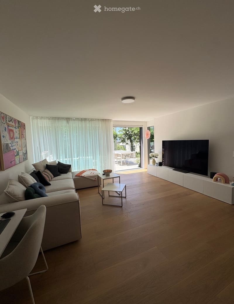 Modern living room with a sectional sofa, coffee table, TV, and large windows leading to a balcony.