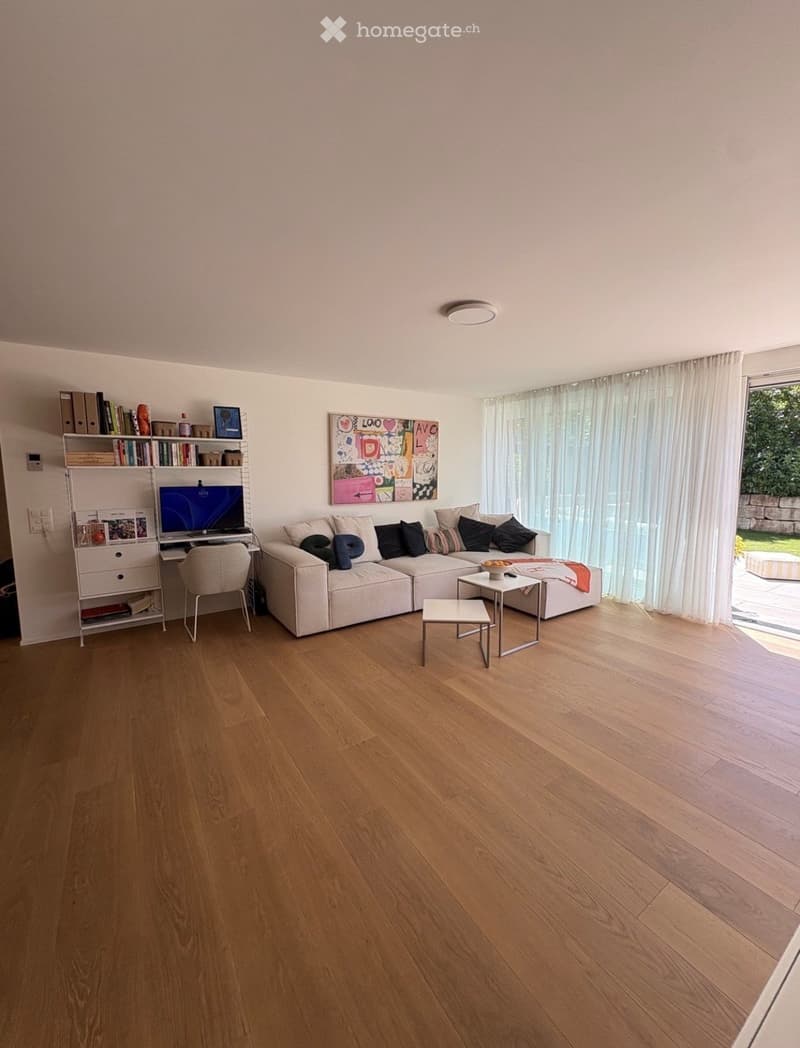 Modern living room with light wooden flooring, a white sectional sofa, a small coffee table, a bookshelf, and a desk area with a screen and chair.