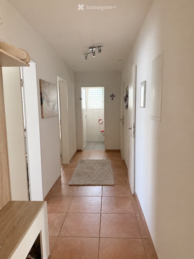 A clean, well-lit hallway with beige tiled flooring, white walls, and a small rug in the center. The hallway features several doors, a small shelf on the left, and a painting on the wall. A bathroom is visible at the end of the hallway.