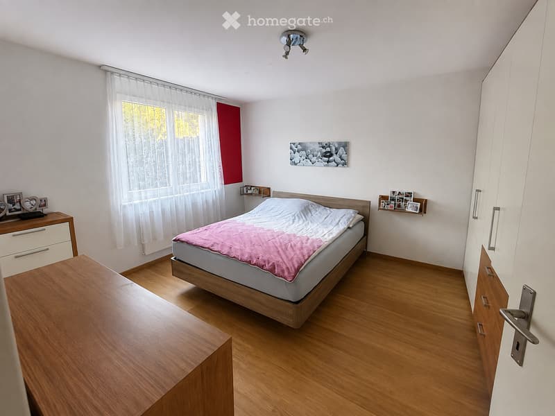 A well-lit bedroom with a wooden bed, pink blanket, nightstand, and a large window with sheer curtains. The room features a wooden floor, white walls, and built-in cabinets.