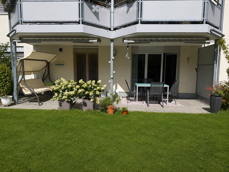 Garden with green lawn, swing chair, small table and chairs, potted plants, and a covered balcony area.