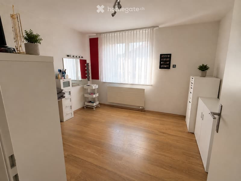 Well-lit room with wooden flooring, large window with white blinds, white walls, and various furniture including a mirror, cabinets, and a small cart.
