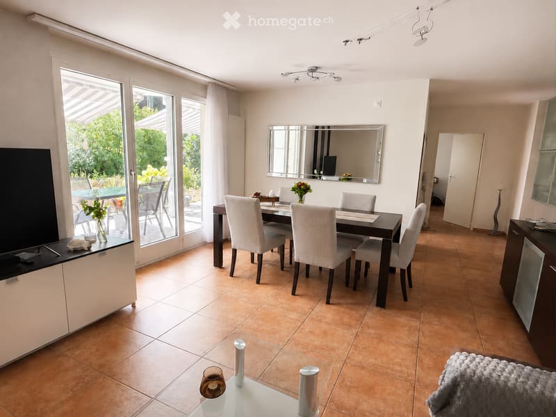 Bright dining area with a dark wooden dining table and six light grey chairs, a large mirror on the wall, a television on a white cabinet, and a glass table with decorative items.