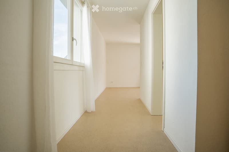Empty hallway with beige carpeting, white walls, and a window with white curtains on the left side.