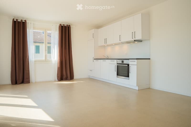 Modern kitchen with white cabinets, stainless steel oven, and a sleek countertop. The space is bright with large windows and brown curtains.