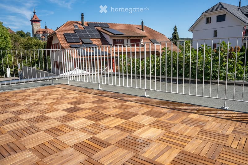 Wooden deck with herringbone pattern, white metal railing, solar panels on adjacent roof, greenery in background.