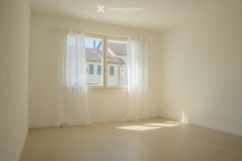 Empty room with light-colored walls, carpeted floor, and large windows with white curtains. No furniture or equipment present.