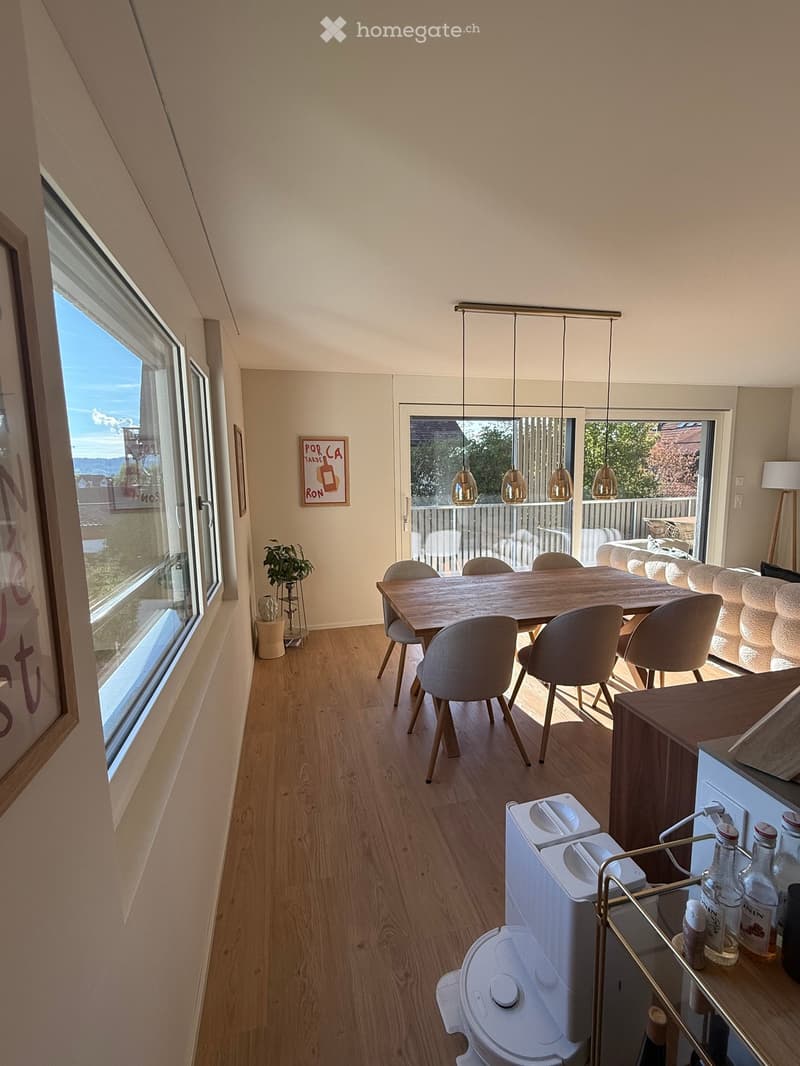 A modern, open-plan living and dining area with wooden flooring, large windows, a dining table with chairs, a sofa, pendant lights, and a small kitchen area with a refrigerator and bottles on the counter.