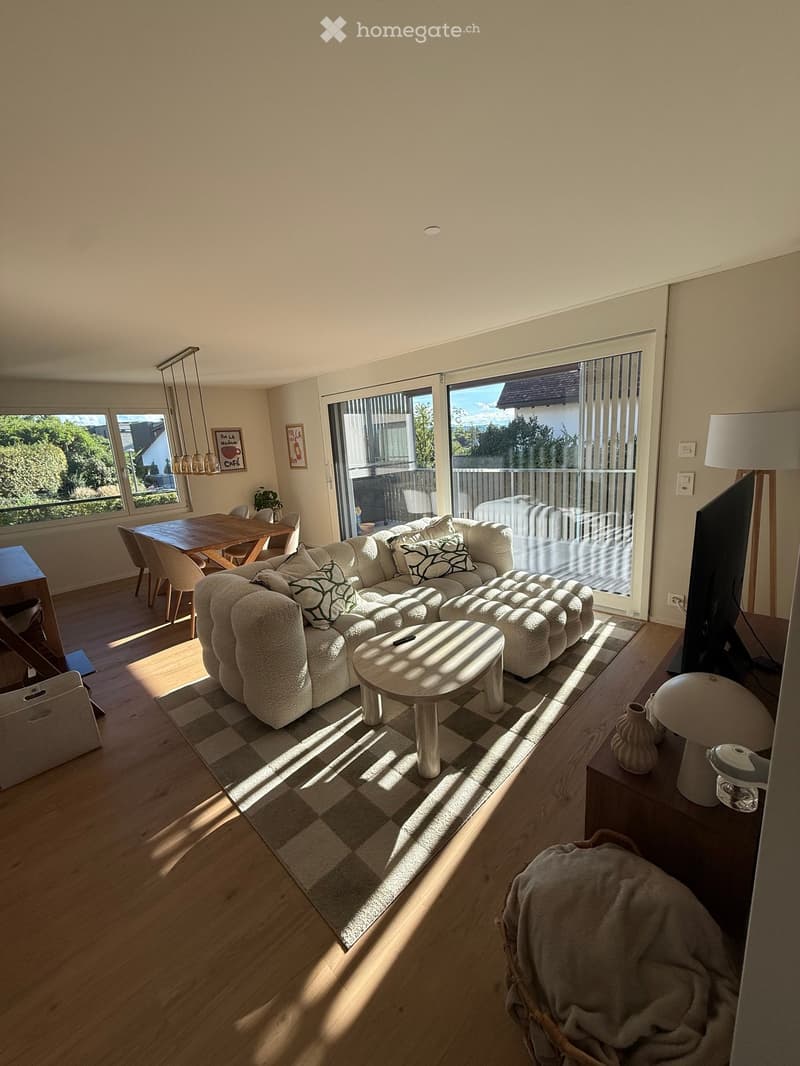 Bright living room with a large sectional sofa, coffee table, dining area, and large windows leading to a balcony.