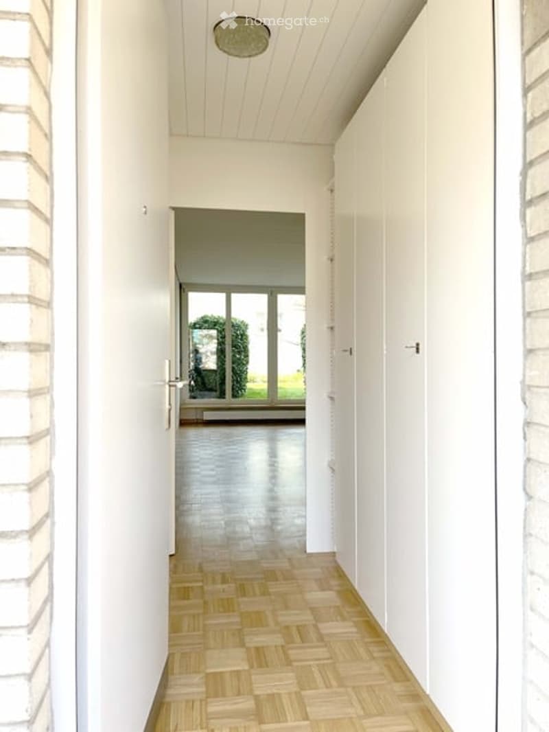 Bright hallway with wooden flooring, white walls, and a ceiling light fixture. The hallway leads to a room with large windows and a view of greenery outside.