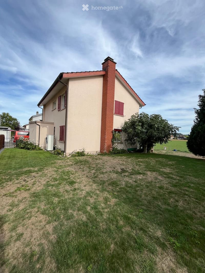 Single-story house with beige walls, red shutters, and a brick chimney. The front yard has a mix of grass and bare patches, with a few trees and a parked van visible in the background.