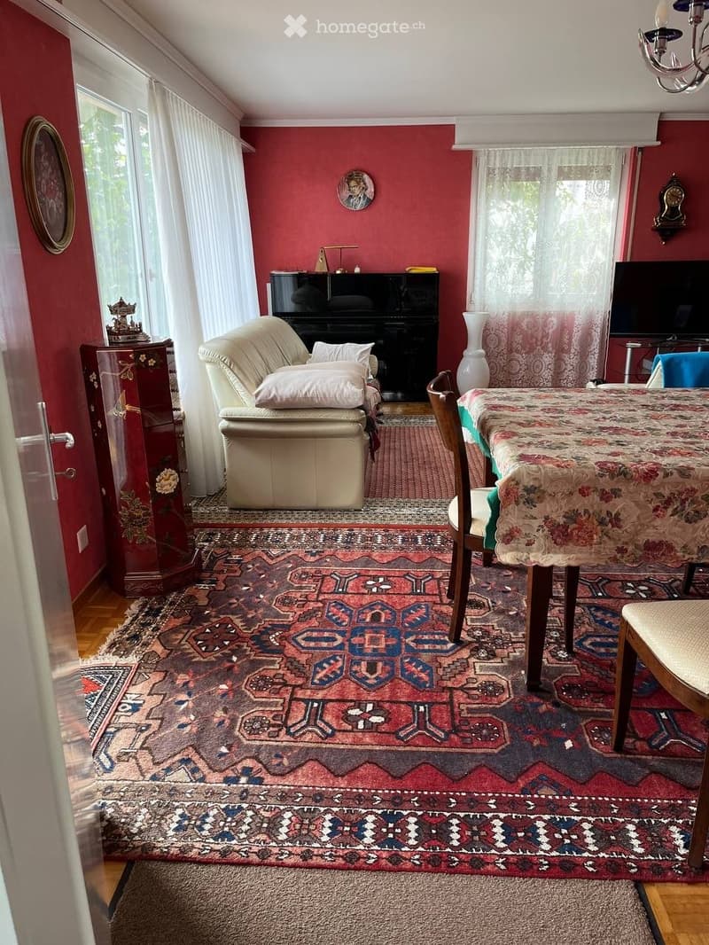 Living room with red walls, large windows, patterned rug, sofa, ottoman, dining table, chairs, piano, television, vases, and a clock on the wall.