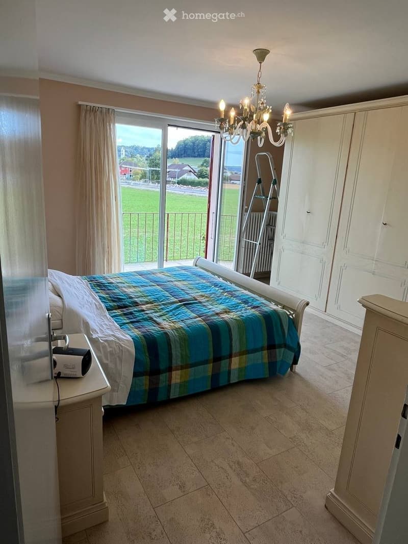 Bright bedroom with a large window offering a scenic view, featuring a bed with a plaid duvet, a nightstand, a chandelier, and built-in wardrobes.