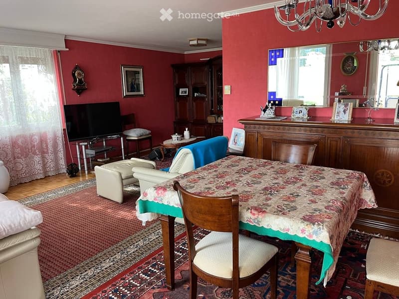 A well-furnished room with red walls, a large mirror, a television, a sofa, a dining table with chairs, and a cabinet with decorative items.