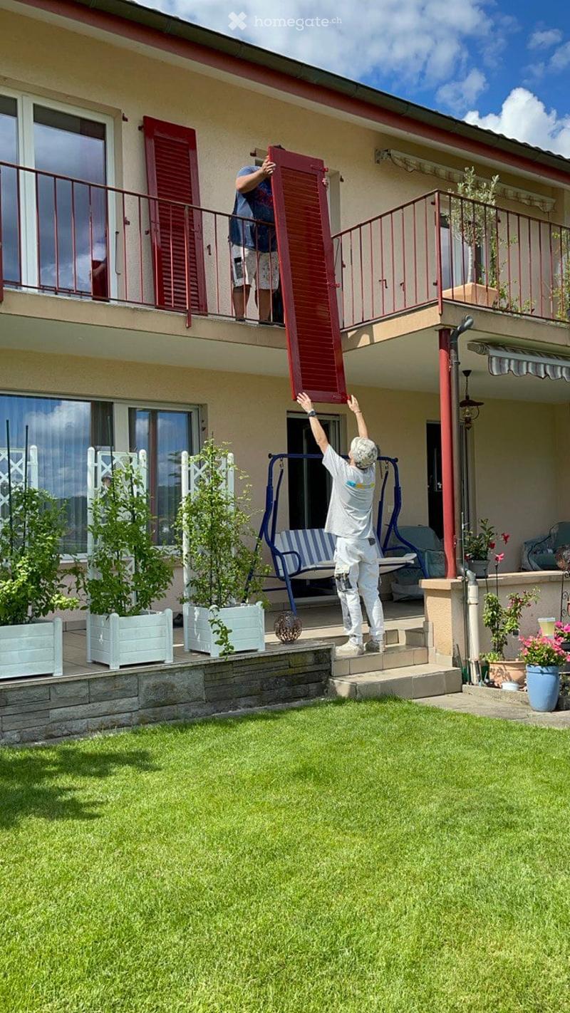 Garden area with green lawn, potted plants, and a swing set. Two men are carrying a large red shutter up the stairs to a balcony.