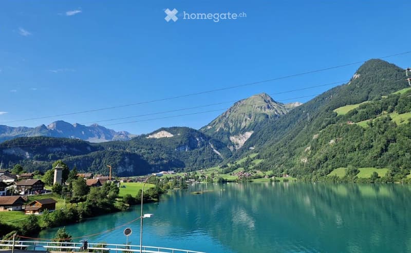 Scenic view of a lake surrounded by lush green hills and mountains, with small houses and a bridge visible in the foreground.