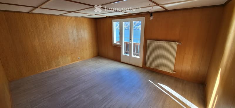 Empty room with wooden walls and floor, large window with a view, radiator, and a hanging light fixture.