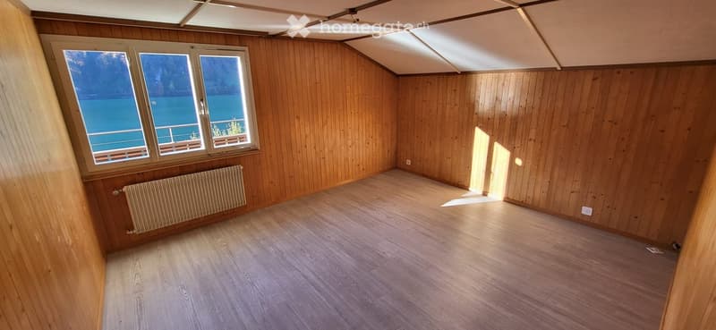 Empty room with wooden walls and floor, large windows overlooking a lake, radiator on one wall.