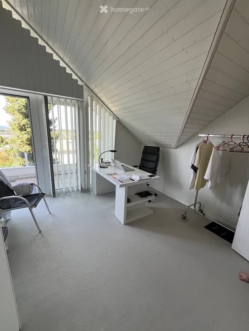 Bright, modern attic office with white walls, slanted wooden ceiling, large windows offering a scenic view, a white desk with a laptop, a black office chair, a floor lamp, and a coat rack with hanging garments.