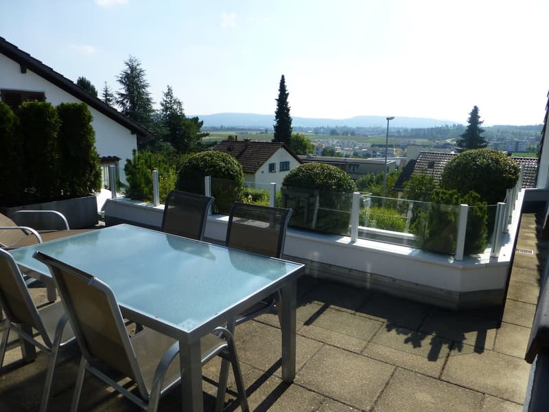 Balcony with a glass table and four chairs, overlooking a scenic landscape with trees and houses.