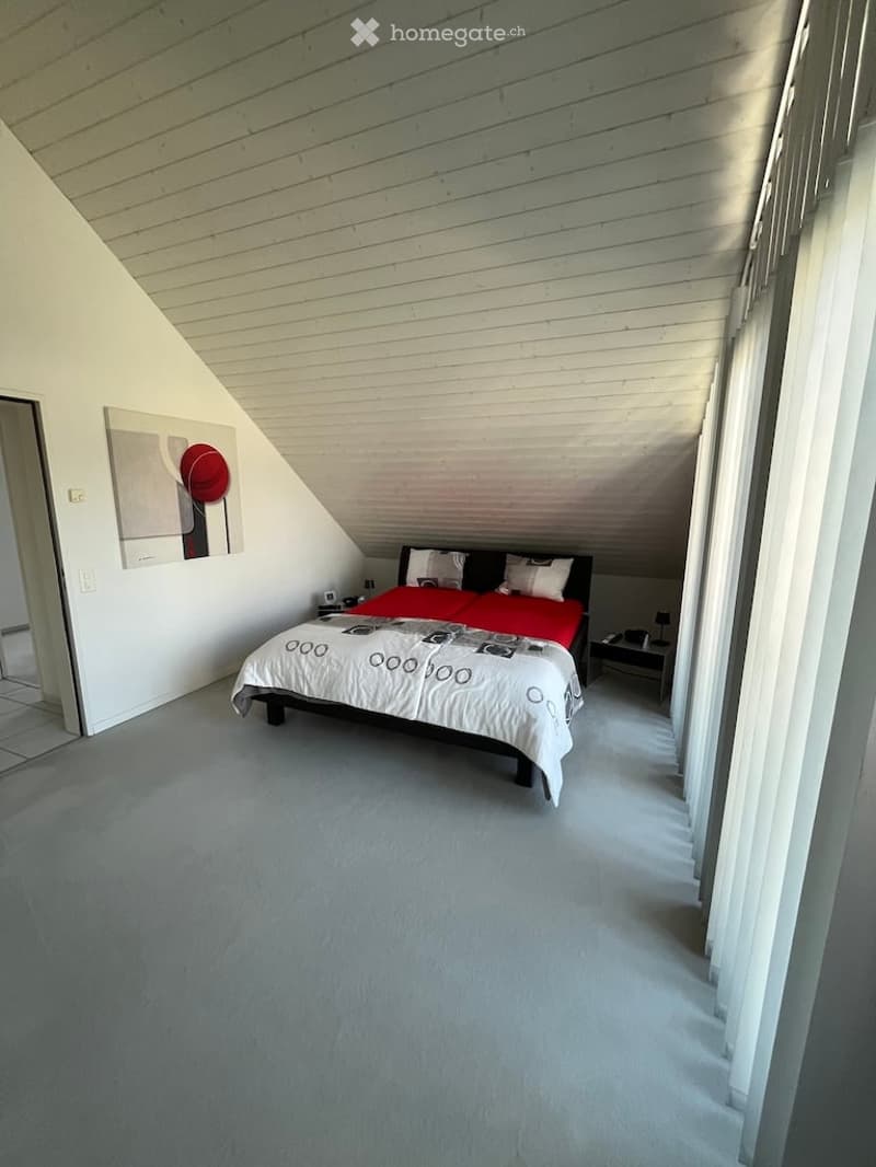 Bedroom with slanted wooden ceiling, grey carpet, white walls, bed with red and white bedding, nightstand, and picture frame on wall.