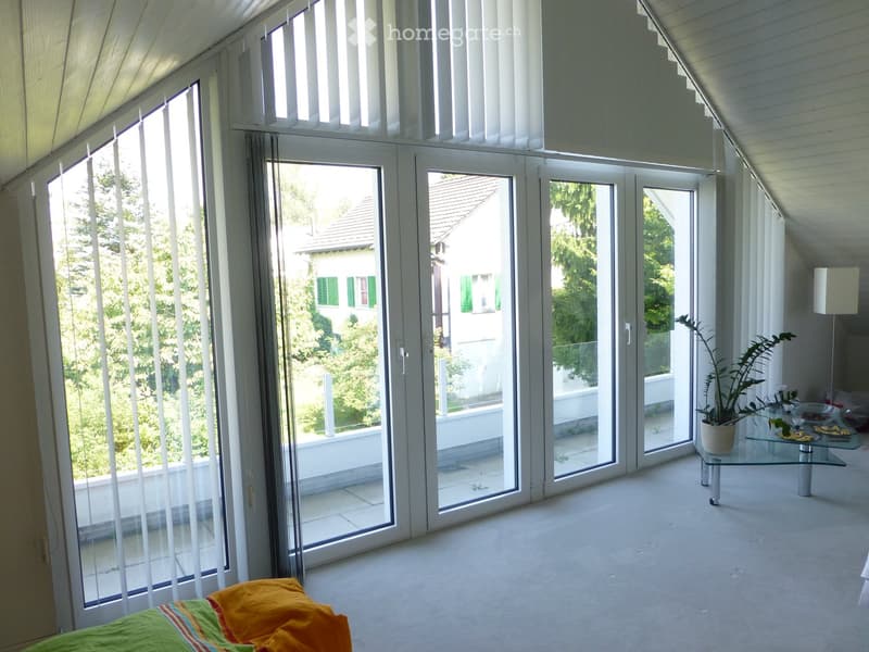 Bright, airy room with large glass doors leading to a balcony, featuring a bed with colorful bedding, a glass table with a potted plant, and a lamp.