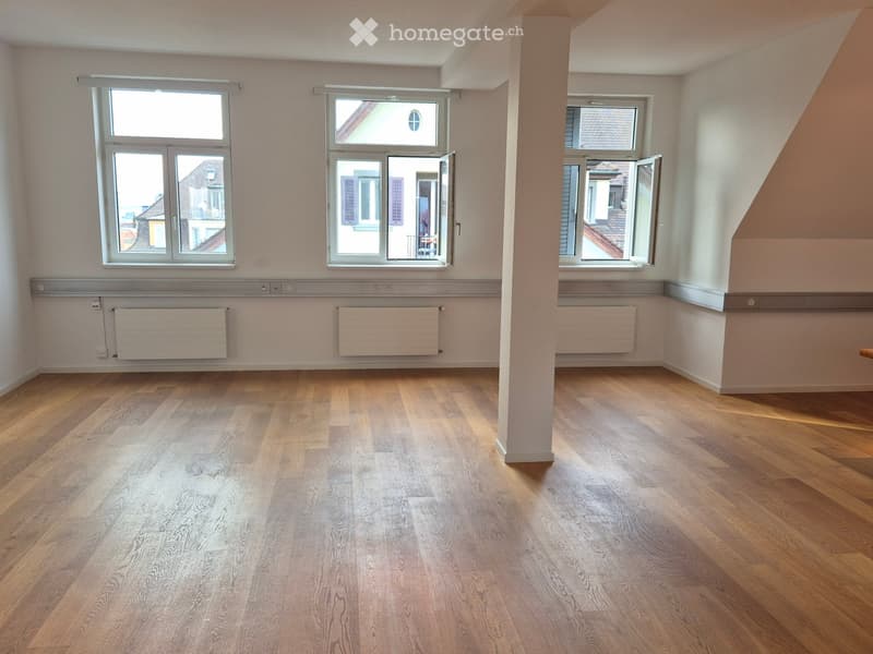 Empty room with wooden flooring, large windows, radiators, and a central column.