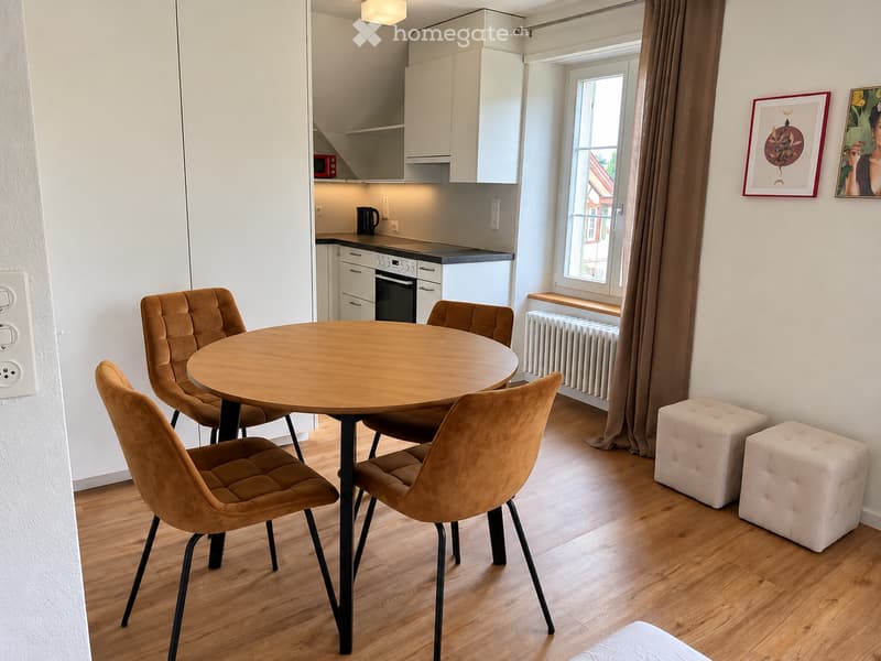 A modern, well-lit room with a round wooden dining table surrounded by four brown upholstered chairs, two white tufted stools, white kitchen cabinets, an oven, microwave, and a window with brown curtains.