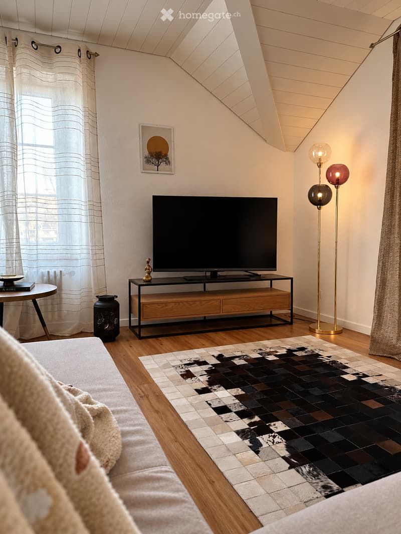 Modern living room with a slanted ceiling, wooden flooring, a large television on a wooden and black stand, a patterned rug, a round side table, and floor lamps.