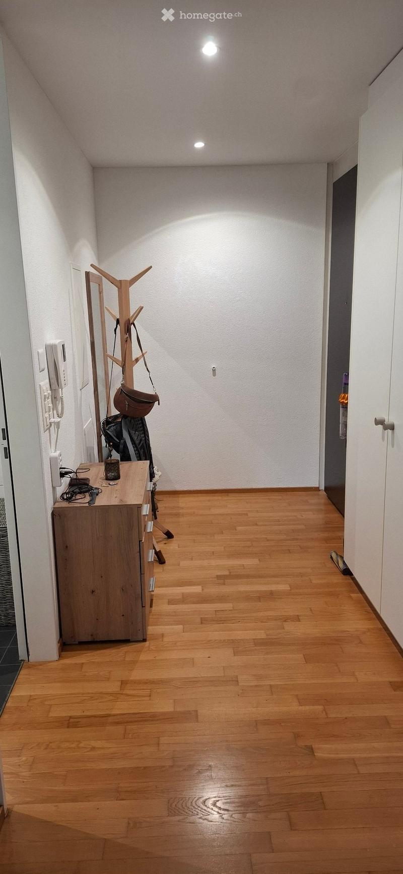 A clean, well-lit hallway with wooden flooring, white walls, and a coat rack with a bag and coat hanging on it. There is a small wooden table with a few items on it, including a cup and some cables.