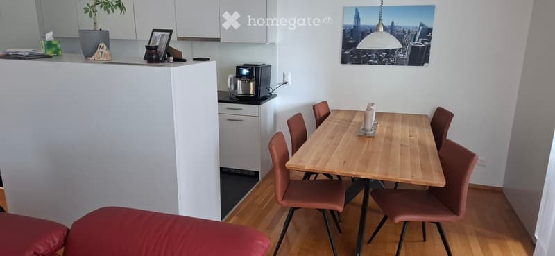 A modern, open-concept living and dining area with a red sofa, wooden dining table, six chairs, a coffee maker, and a potted plant on the counter.