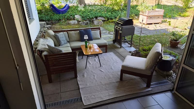 Balcony with a sofa, small table, and chair, surrounded by greenery and a hammock in the background.