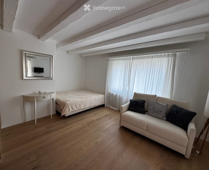 Cozy attic room with wooden flooring, a single bed, a sofa, a small table, and a mirror with a TV mounted on it.