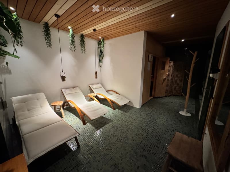 A cozy, well-lit relaxation area with a wooden ceiling, white walls, and a tiled floor. Features include three white chaise lounges, small wooden stools, and hanging plants.