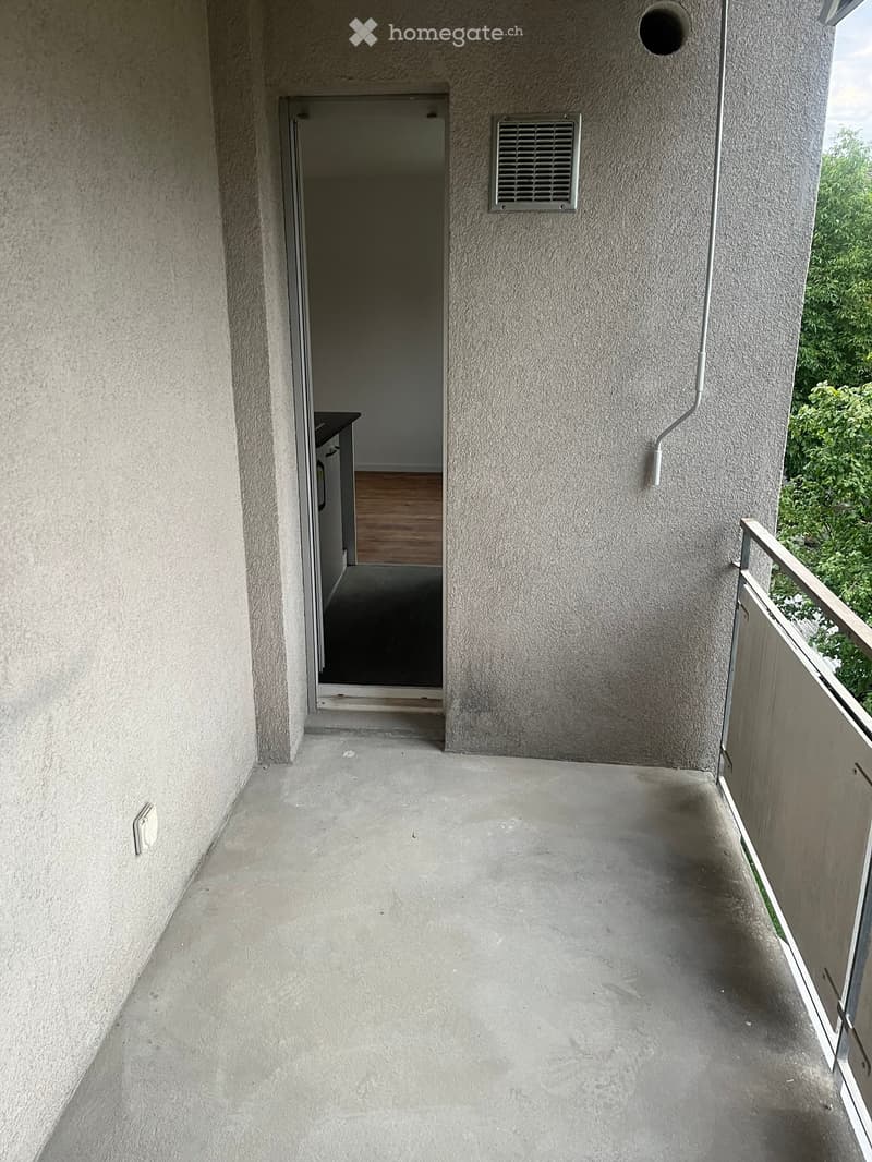 Empty balcony with concrete floor, metal railing, and an open door leading to an interior space.