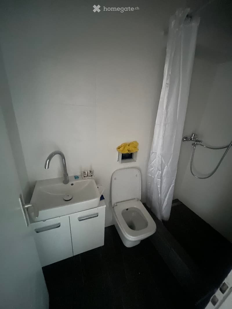 Small bathroom with a white sink, faucet, toilet, and a shower curtain. The floor is dark, and the walls are white.
