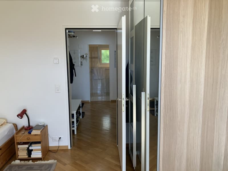 A well-lit hallway with wooden flooring, leading to a bedroom and a bathroom. The hallway features a small wooden shelf with shoes, a red desk lamp, and a nightstand with books and a phone.