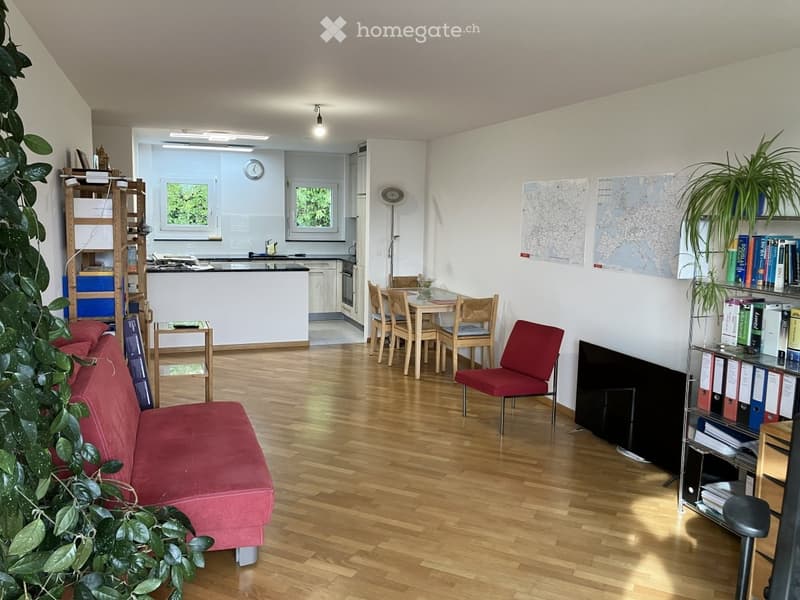 Open-plan living and dining area with wooden flooring, a red sofa, a dining table with chairs, a bookshelf, and a small kitchen area with a counter and oven.