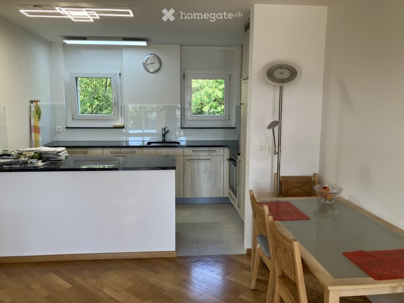 Modern kitchen and dining area with wooden flooring, white cabinets, a black countertop, a dining table with chairs, and a floor lamp.