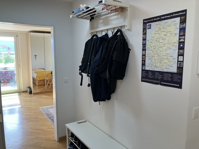 Hallway with wooden flooring, coat rack with three backpacks, a map poster, a small white table with shoes, and a mirror leading to a bedroom area.