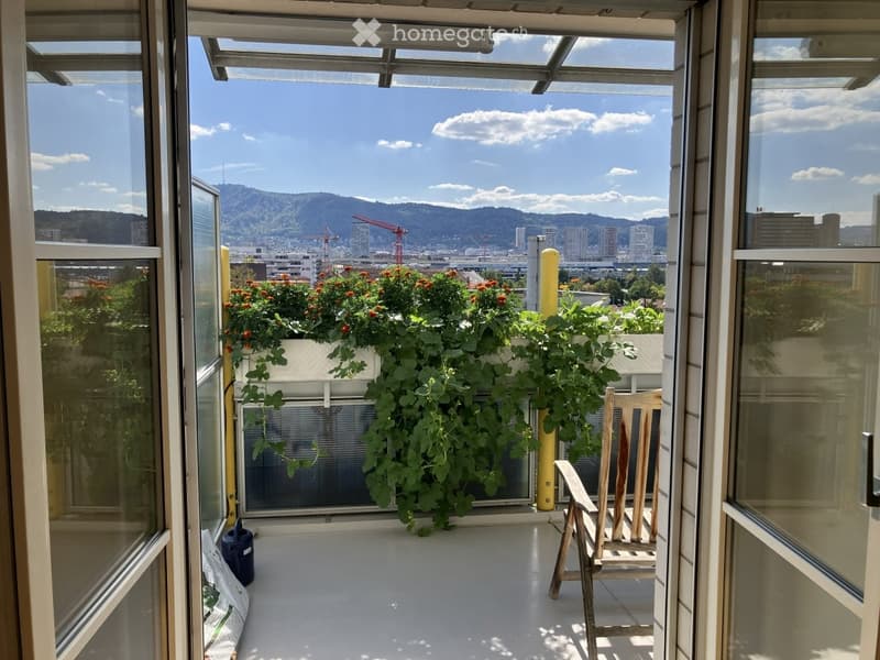 Balcony with a wooden chair, a planter with green plants, and a scenic city view with mountains in the background.