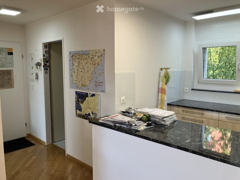 Bright kitchen with white walls, wooden flooring, granite countertop, and two maps on the wall. A towel hangs on a rack, and a doorway leads to another room.