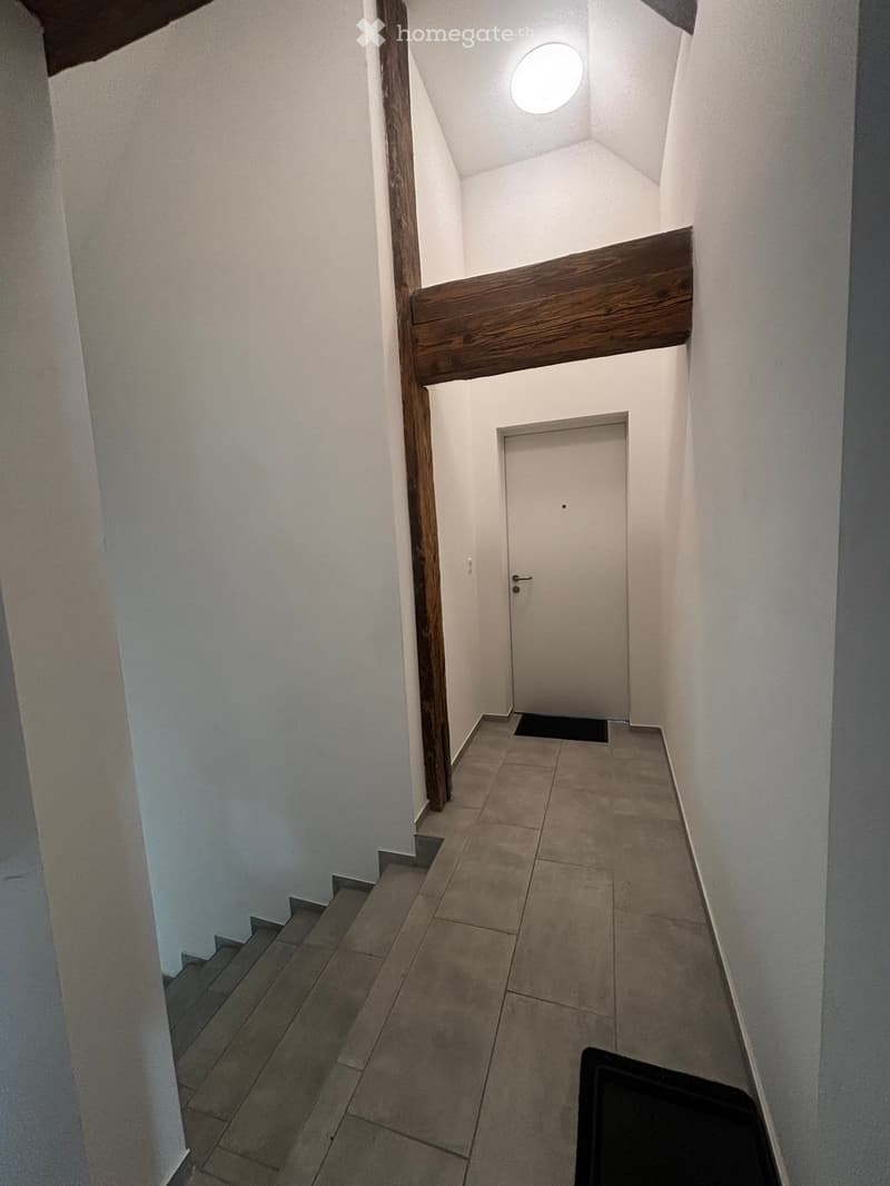 A narrow hallway with white walls, tiled floor, and a wooden staircase leading upwards. A closed door is visible at the end of the hallway.