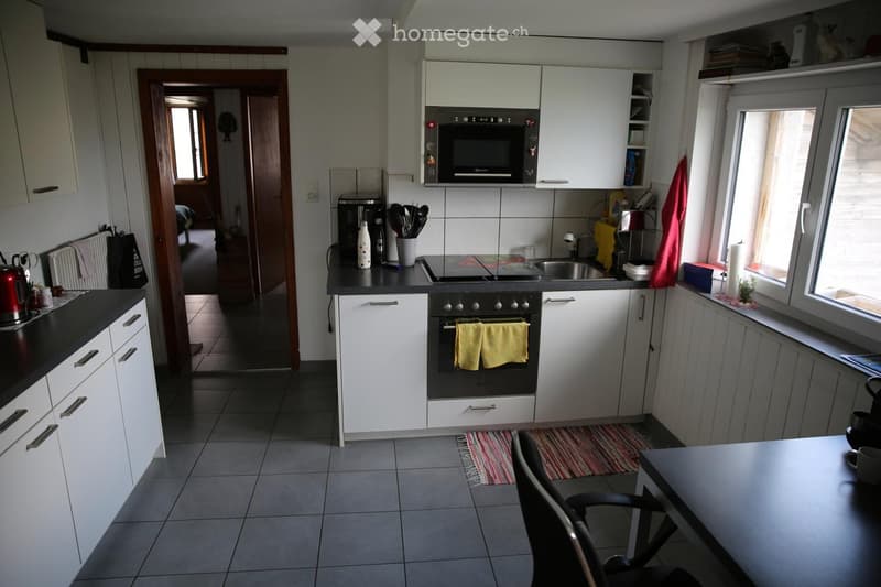 Modern kitchen with white cabinets, black countertops, and stainless steel appliances including an oven, microwave, and sink. A small table with a chair is visible, along with various kitchen utensils and a striped rug on the floor.