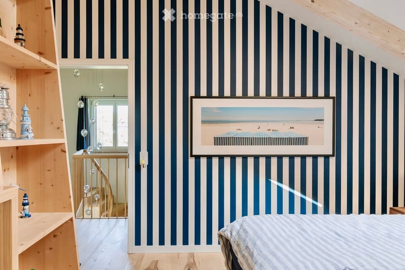 Cozy bedroom with blue and white striped walls, wooden floor, bed with striped bedding, shelf with decorative items, framed beach scene picture, and a staircase leading to another level.