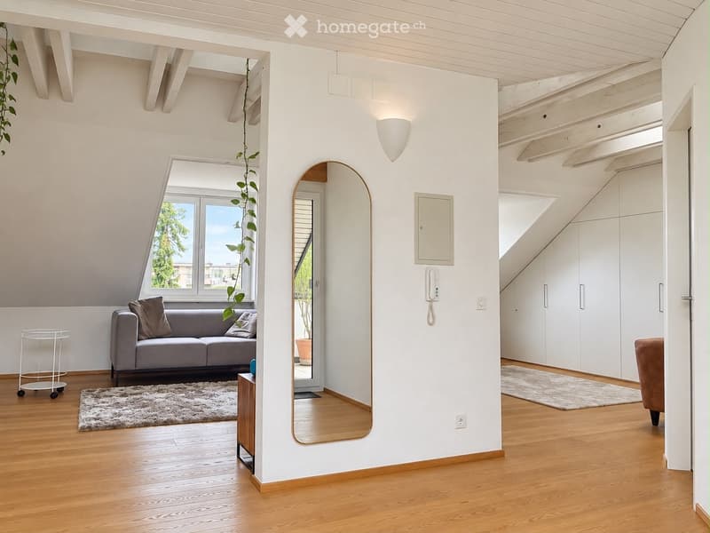 Bright, modern living space with wooden flooring, white walls, and exposed wooden beams. Features a grey sofa with cushions, a round mirror, a side table, a small cart, and a potted plant near the window.
