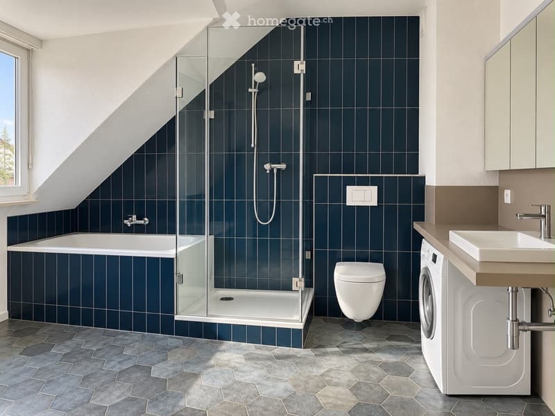 Modern bathroom with dark blue tiled walls, a bathtub, a glass-enclosed shower, a toilet, a sink, and a washing machine. The room features hexagonal gray tiles on the floor and a window with a view of greenery.