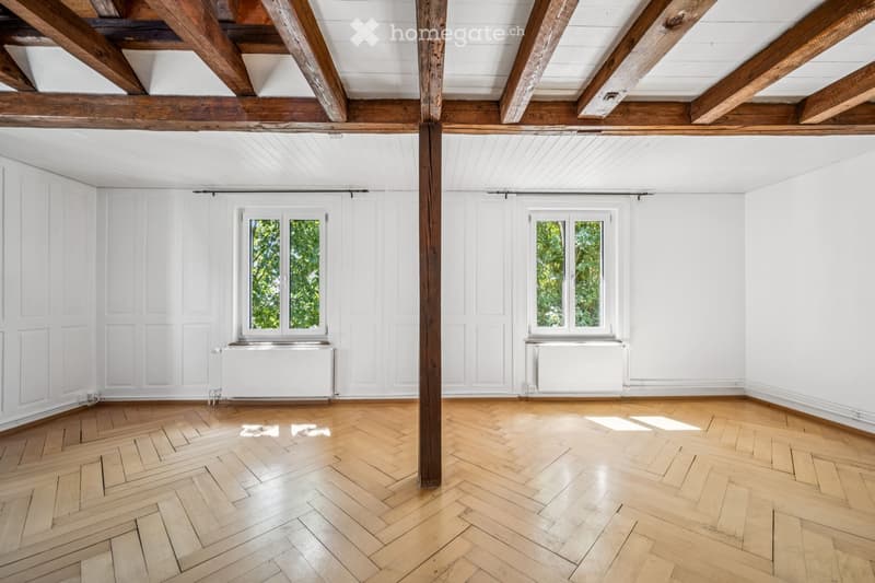 Empty room with wooden herringbone flooring, white walls, exposed wooden beams, and two windows with white frames.