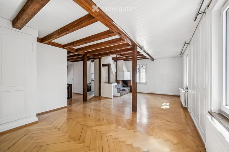 Bright, open corridor with exposed wooden beams and herringbone parquet flooring. White walls, large windows, and visible kitchen area in the background.